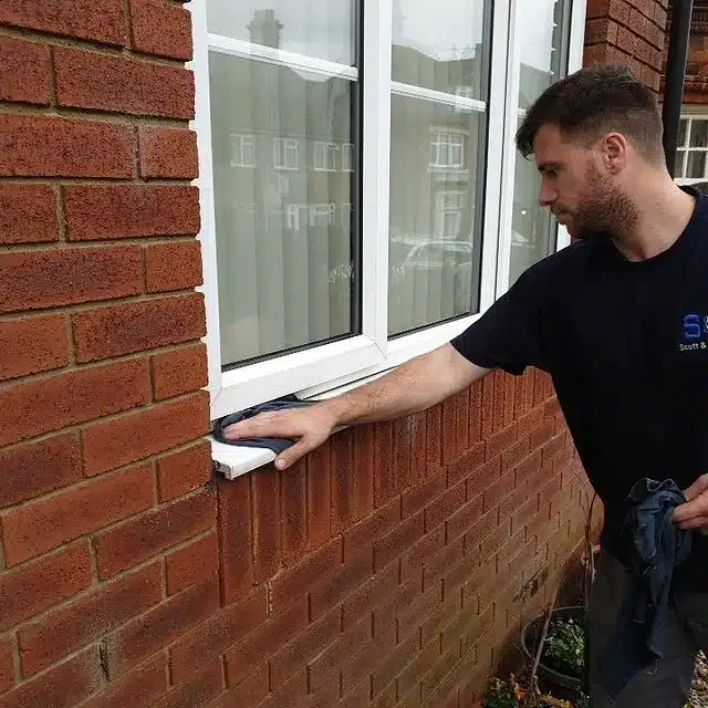 Scott & Sons team member cleaning outdoor window frame seals on a brick building as part of a spring cleaning.