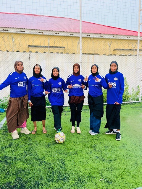 Six young women in blue soccer uniforms and hijabs, supported by kit aid from Scott & Sons, stand on an outdoor soccer field, posing for a group photo with a soccer ball in front of them.